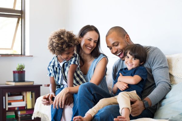 Family having fun at home Happy multiethnic family sitting on sofa laughing together. Cheerful parents playing with their sons at home. Black father tickles his little boy while the mother and the brother smile.