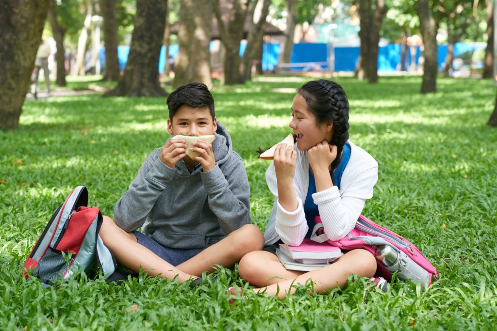 Happy school kids sitting on grass in park eating sandwiches for lunch